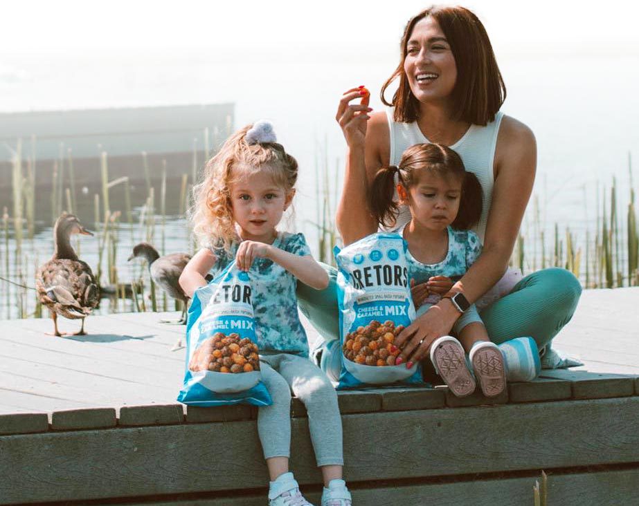 Mom sitting on a dock with two little girls, eating from a bag of caramel and cheese popcorn while ducks waddle in the background. 