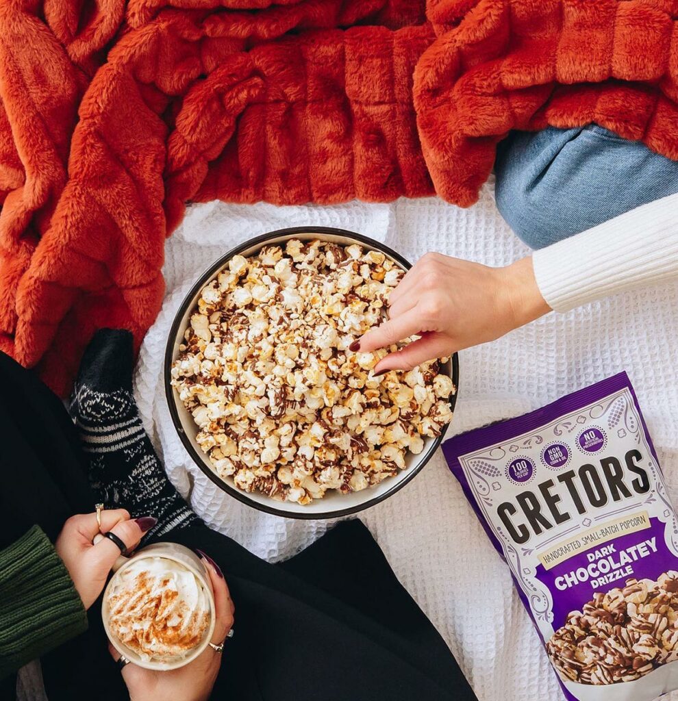 Cozy overhead image of a woman’s hands holding a mug of hot chocolate and another woman’s hand selecting a piece of dark chocolate drizzle Cretors popcorn from a bowl next to a soft blanket. 