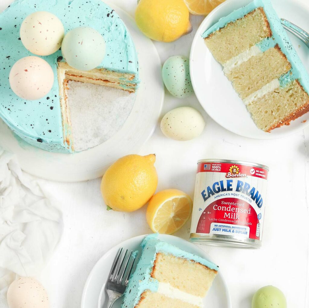 Can of Eagle Brand Sweetened Condensed Milk lying among a sliced cake with blue icing, eggs and cut lemons. 