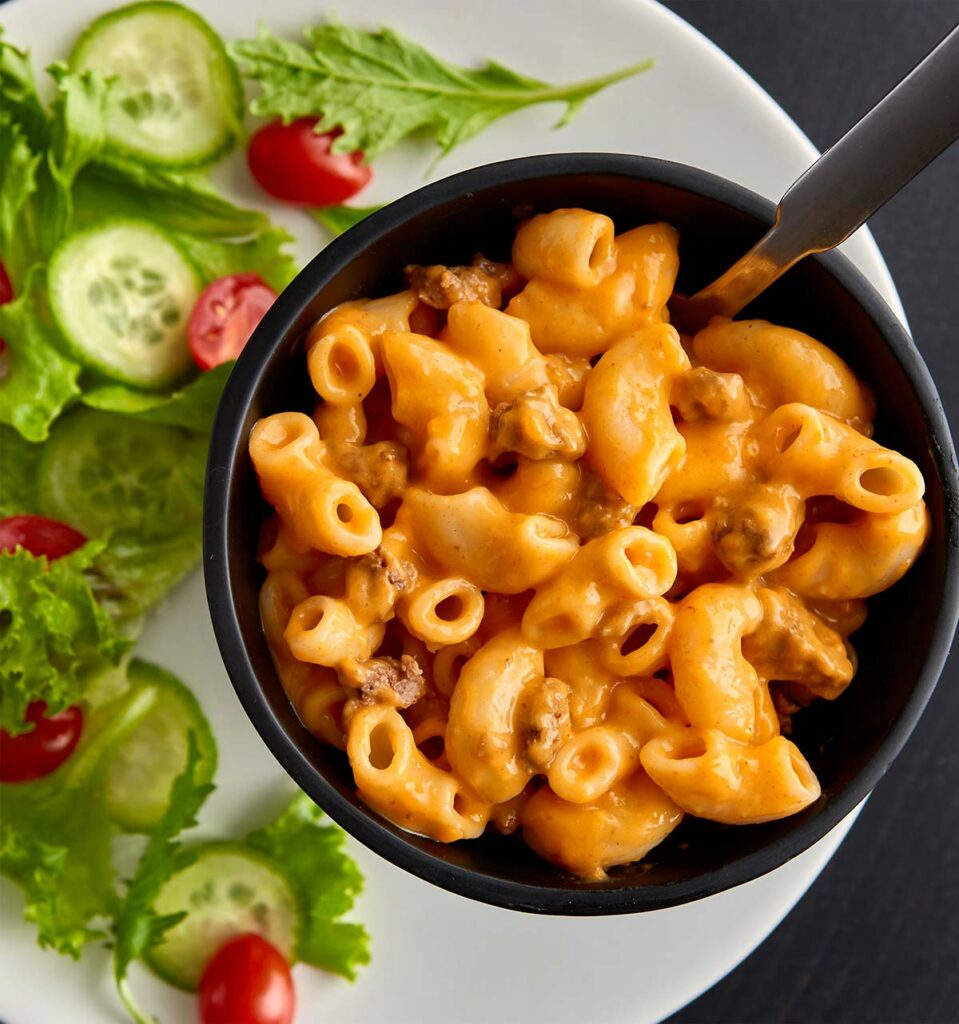 Black bowl of creamy Hamburger Helper noodles next to a leafy green side salad with tomatoes and cucumbers. 