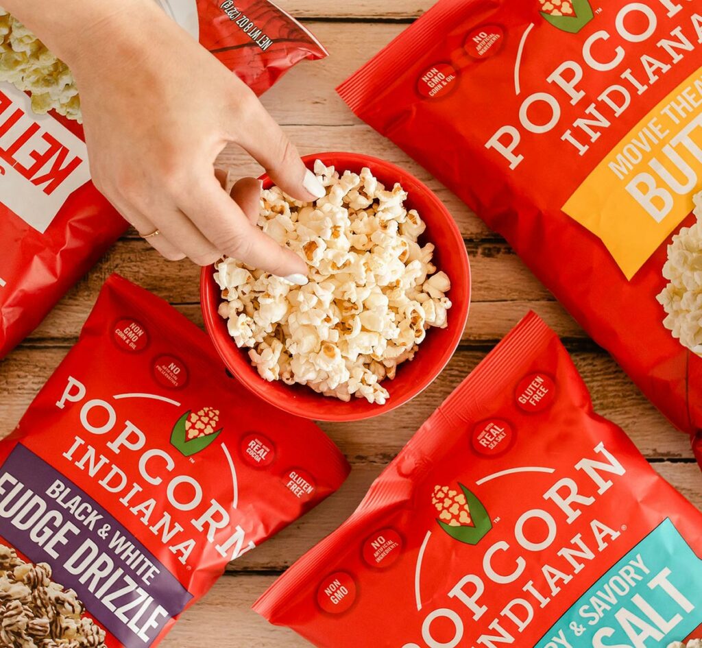 Overhead view of a woman’s hand selecting a piece of popcorn from a red bowl nestled in the center of four red bags of Popcorn, Indiana.