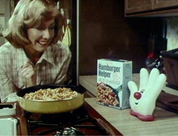 Blonde woman smiling as she overlooks a cooking pan full of Hamburger Helper with the product box sitting on the counter next to Lefty, the white glove “spokes character.” 