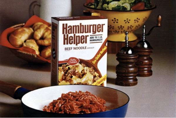 Box of Hamburger Helper featured next to a blue pan full of Helper and dinner rolls with salt and pepper shakers in the background. 
