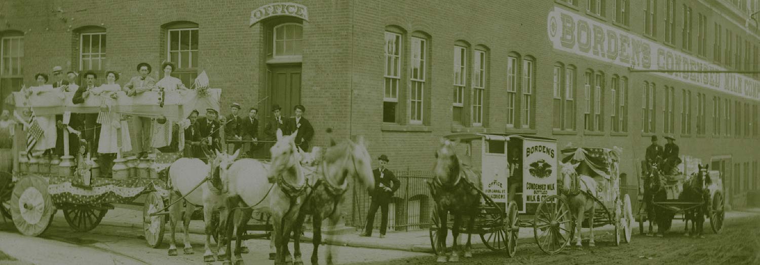 Old-time black and white photo of horse-drawn carriages parked in front of the Borden’s Condensed Milk factory. 