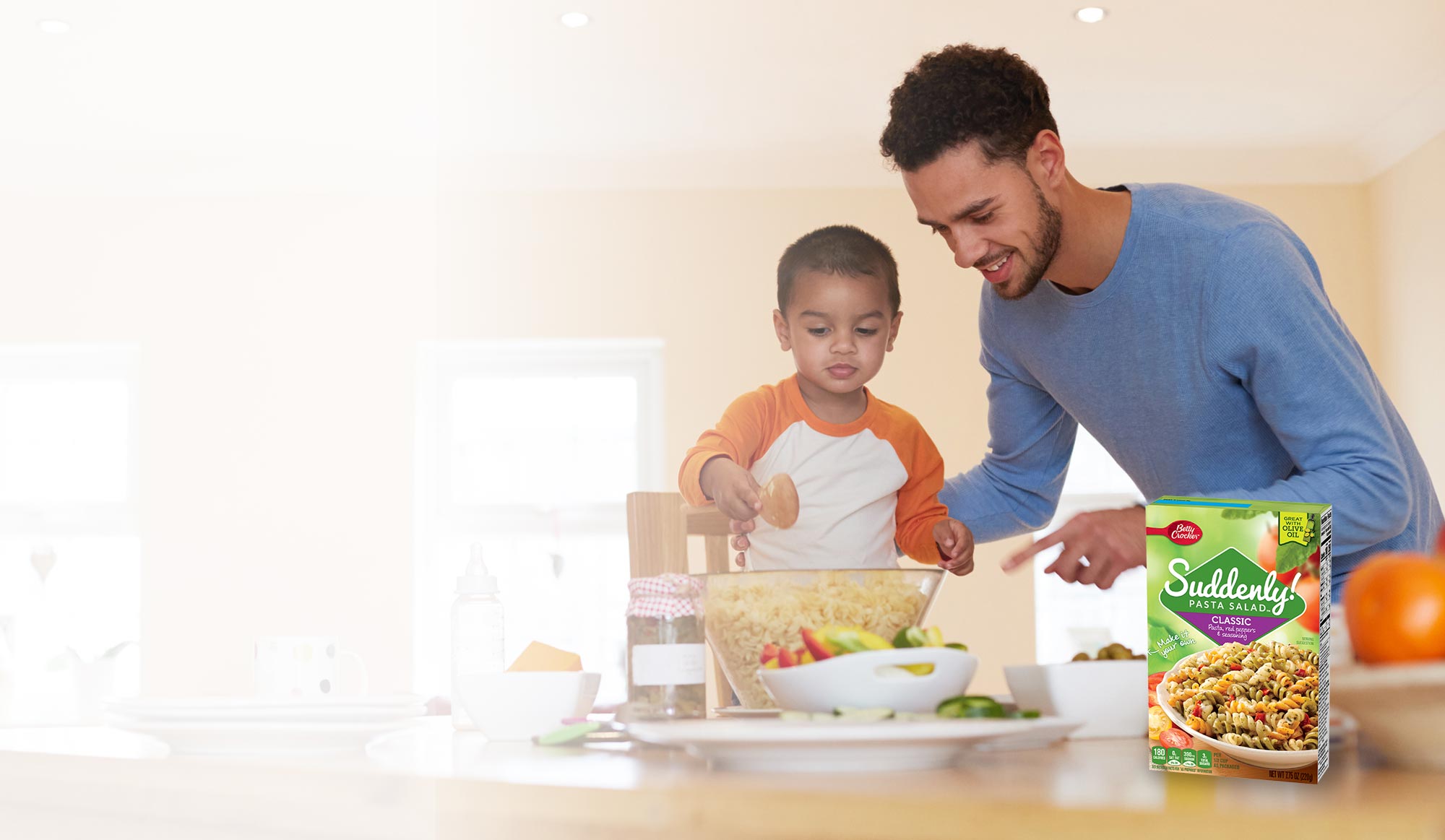 Father helps his young son in the kitchen stir and make some Suddenly Salad pasta salad, with product box displayed on the counter.