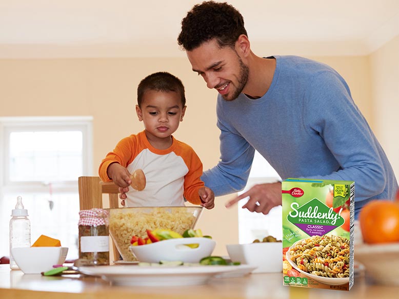 Father helps his young son in the kitchen stir and make some Suddenly Salad pasta salad, with product box displayed on the counter.