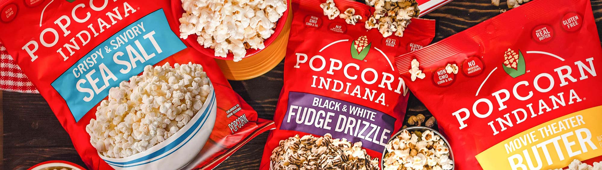 Overhead view of tabletop with three red bags of Popcorn Indiana products: Crispy & Savory Sea Salt on the left, Black & White Fudge Drizzle in the middle, and Movie Theater Butter on the right. 