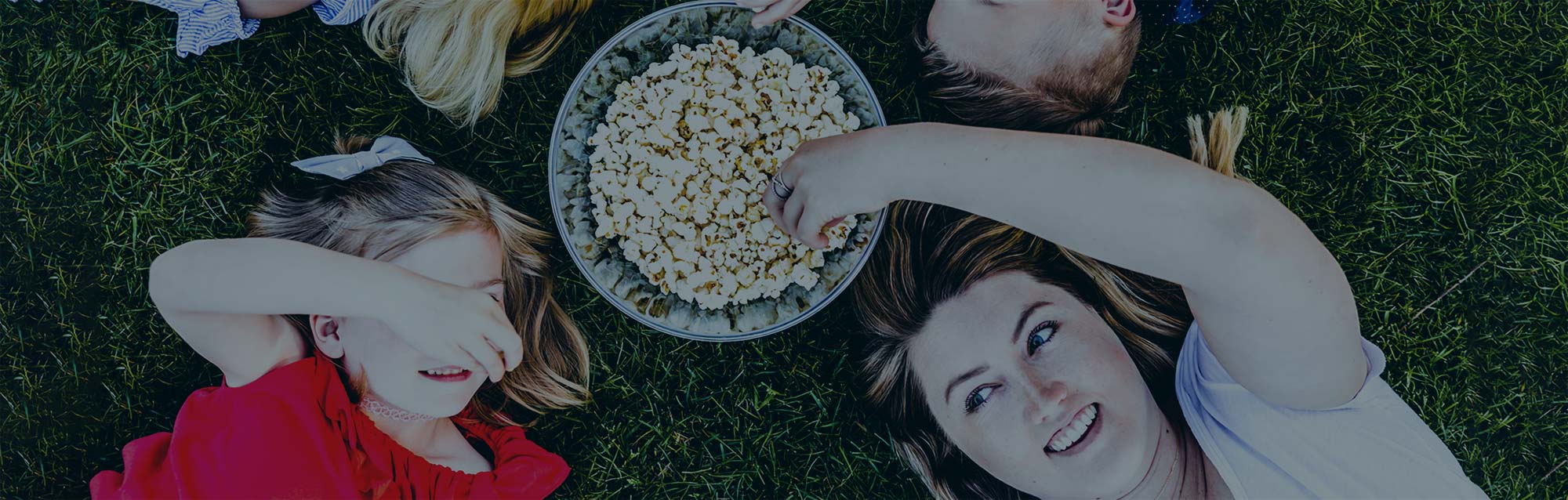 Children lying on their backs in a circle on the grass reach overhead and into a bowl of popcorn that is between them.