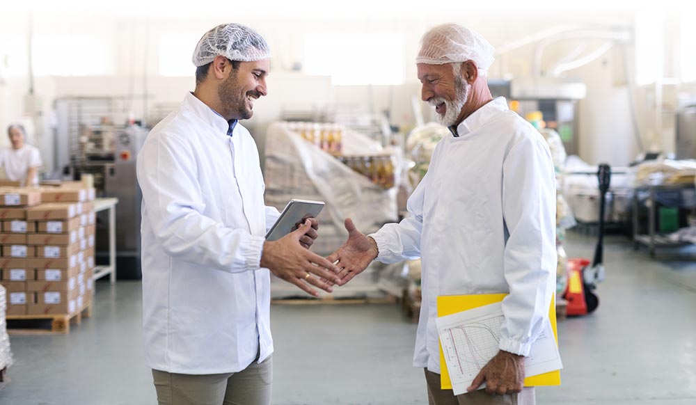 Two male food inspectors wearing white lab coats and hair nets smile and greet one another with a hearty handshake.