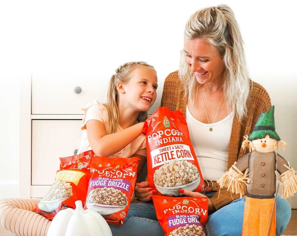 Mother and daughter sitting side-by-side, smiling and sampling multiple red bags of various Indiana Popcorn flavors.