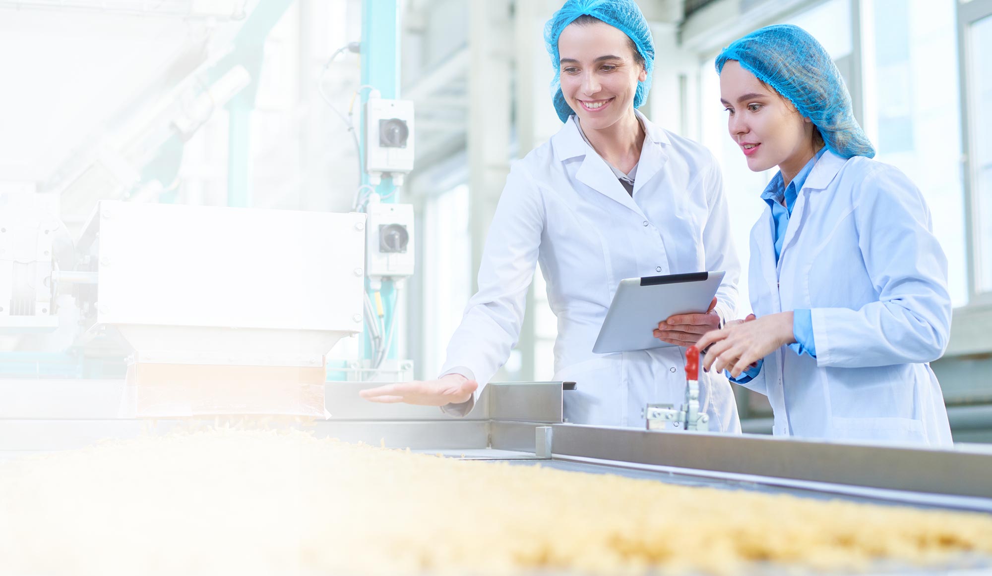 Two female food inspectors wearing white lab coats and blue hair nets smile as they look over a conveyor belt of food.