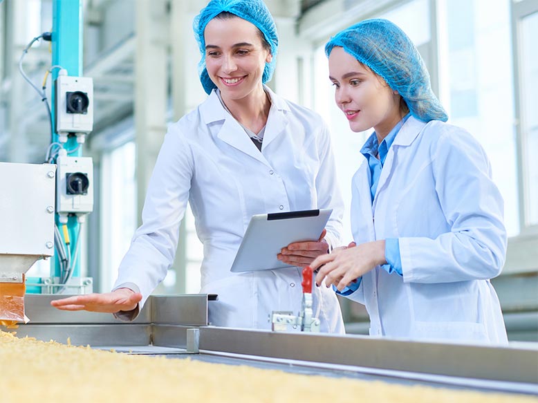 Two female food inspectors wearing white lab coats and blue hair nets smile as they look over a conveyor belt of food.